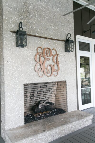A textured stone outdoor fireplace with a black brick interior, flanked by two black lanterns. A decorative metal monogram hangs above the opening, and glass doors are visible to the right.