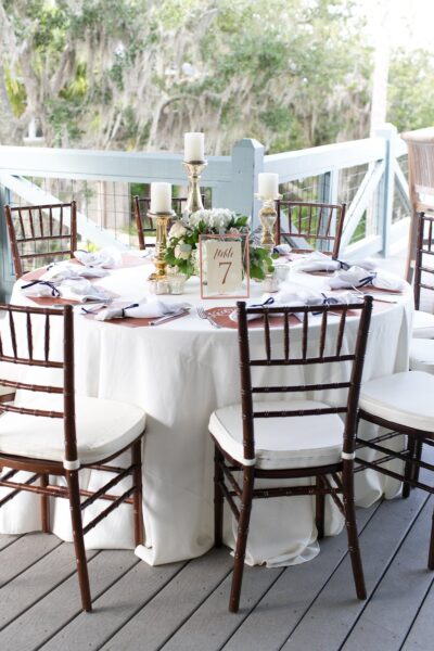 A round table with a white tablecloth is set for six, featuring dark wooden chairs, white napkins, gold candlesticks, a floral centerpiece, and a table number card reading 7 on a wooden deck outdoors.