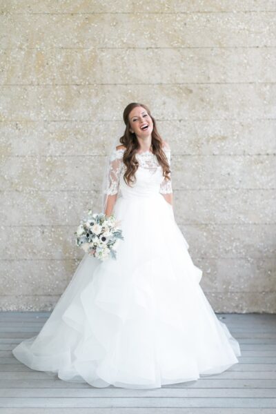 A bride in a white, off-the-shoulder lace wedding dress stands smiling and laughing, holding a white and green bouquet, in front of a light beige textured wall.