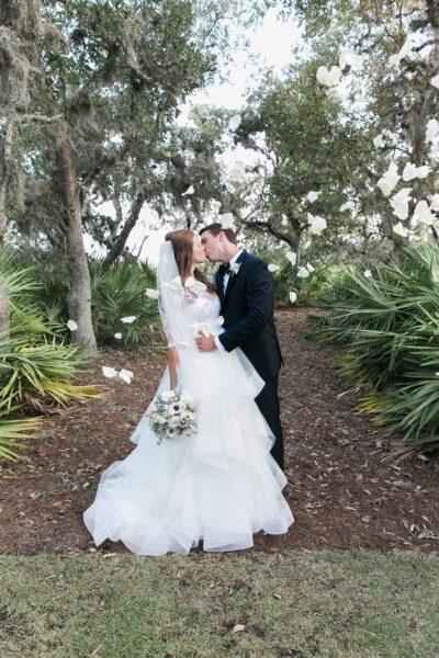 A bride and groom share a kiss outdoors on a tree-lined path. The bride wears a white gown and holds a bouquet, while the groom wears a black suit. White flower petals float around them.