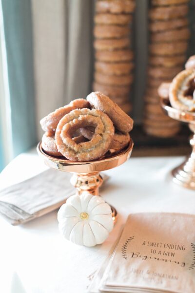 Glazed donuts arranged on a gold stand, with stacked donuts in the background; a small white pumpkin and personalized napkins reading “A sweet ending to a new beginning” are displayed on the table.