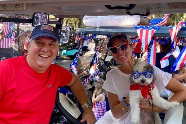 A smiling man and woman decked in patriotic outfits pose on a decorated golf cart for a Fourth of July celebration. The woman holds a small dog wearing blue star-shaped glasses and a red scarf. American flags and festive decorations are visible.