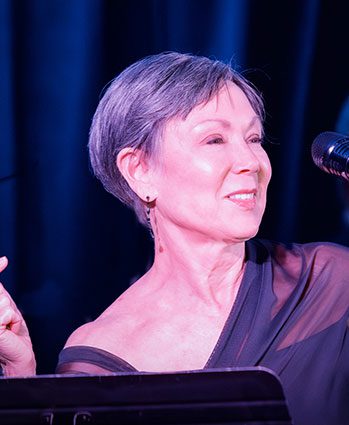 A woman with short gray hair smiles while holding a pointer near a microphone, wearing an off-shoulder dress on stage with dark blue lighting in the background.