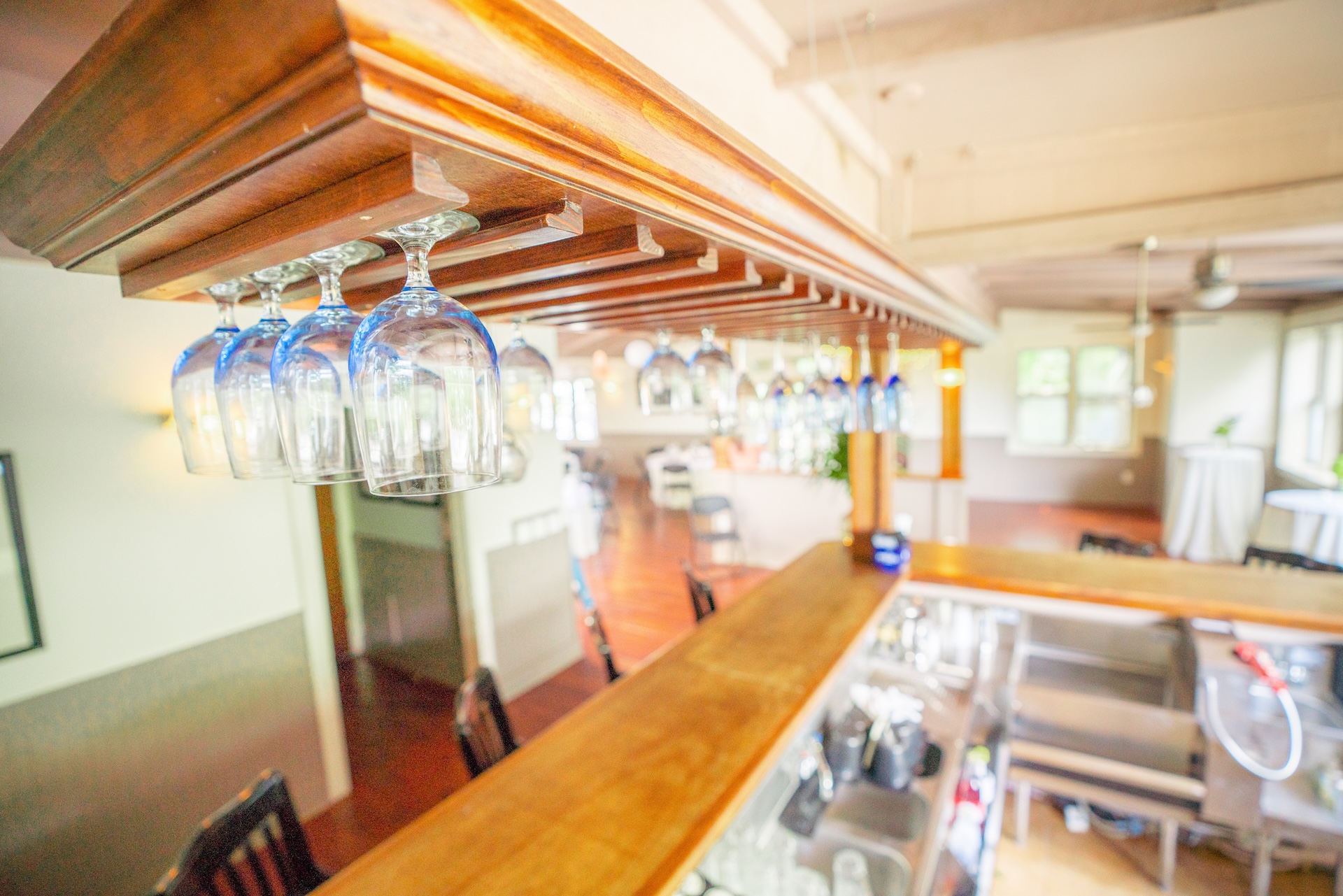 A row of upside-down wine glasses hangs from a wooden rack above a bar in a bright, modern restaurant with wooden floors and white walls.