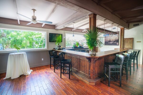 A cozy bar area with wooden floors, a wooden bar counter, black chairs, large windows with greenery outside, and vases with palm leaves on the counter and a nearby round table.