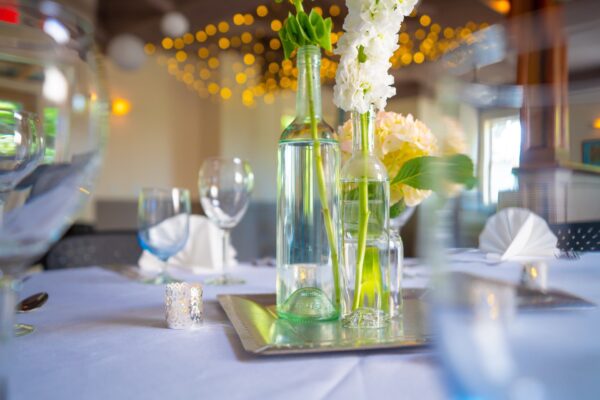 A close-up of a decorated table set for an event, featuring glass bottles with flowers, folded white napkins, and wine glasses on a white tablecloth, with string lights blurred in the background.