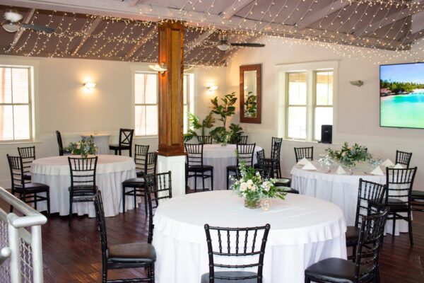 A bright, elegant event room with round tables covered in white cloths, black chairs, floral centerpieces, string lights on the ceiling, and a large TV displaying a beach scene on the wall.