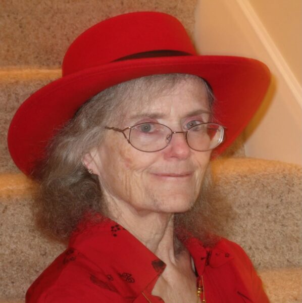 An older woman with gray hair, glasses, and a red wide-brimmed hat sits on carpeted stairs. She is wearing a red shirt and is smiling gently at the camera.