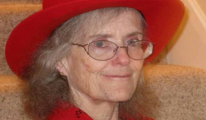 An older woman with gray hair, glasses, and a red wide-brimmed hat sits on carpeted stairs. She is wearing a red shirt and is smiling gently at the camera.