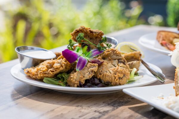 A plate of crispy fried mushrooms garnished with fresh greens and edible flowers, served with a small cup of dipping sauce and a lemon wedge on a wooden table.