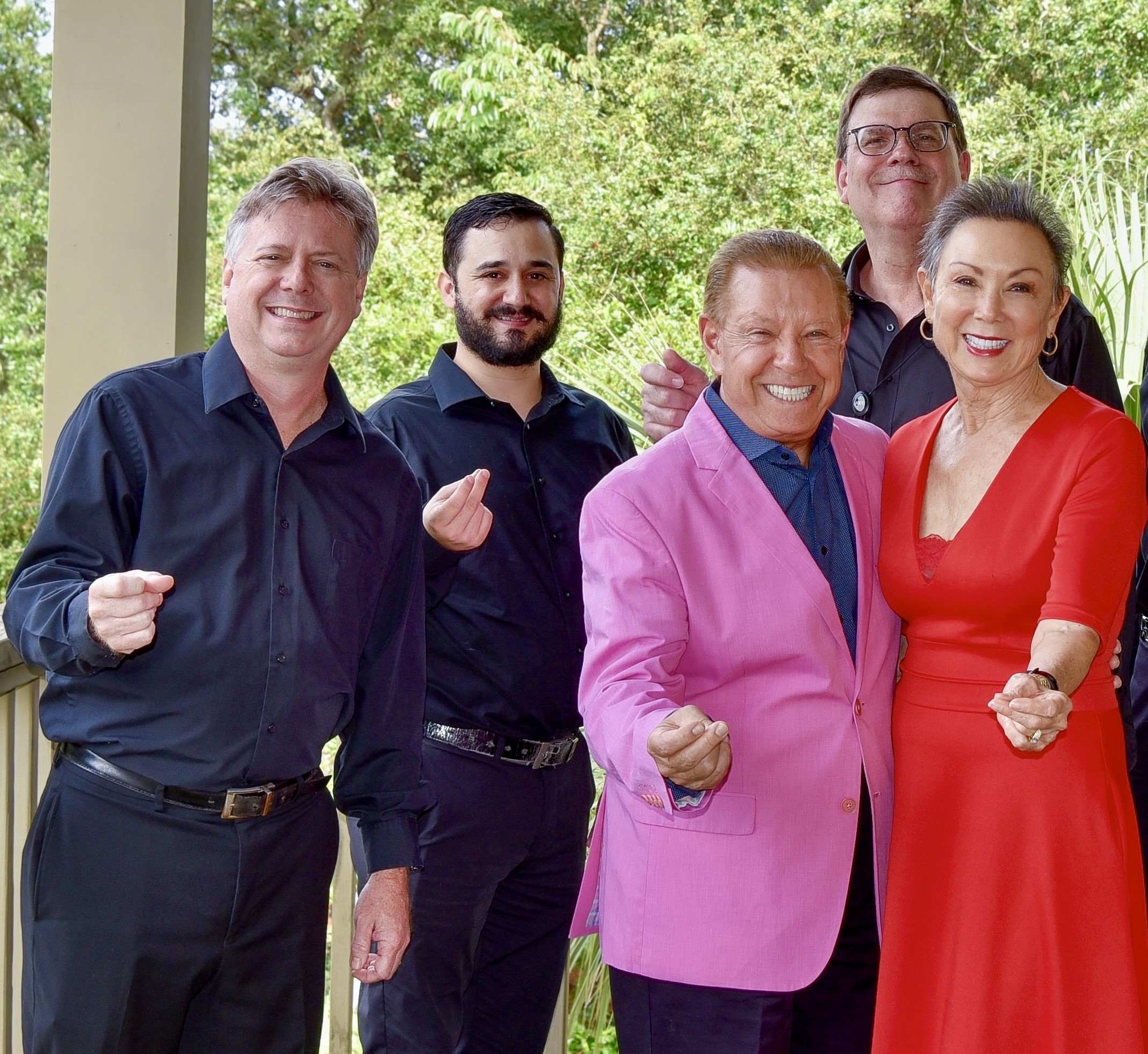 Five adults stand together smiling on a porch. Four men wear black shirts, except one in a bright pink jacket. A woman in a red dress stands in front. They gesture with their fingers as if pinching or holding something small. Greenery is behind them.