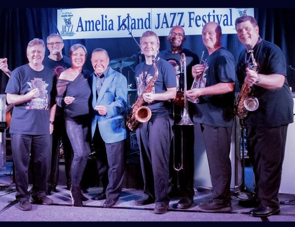 A group of musicians and singers pose together on stage, holding instruments and smiling, with an Amelia Island Jazz Festival banner in the background.