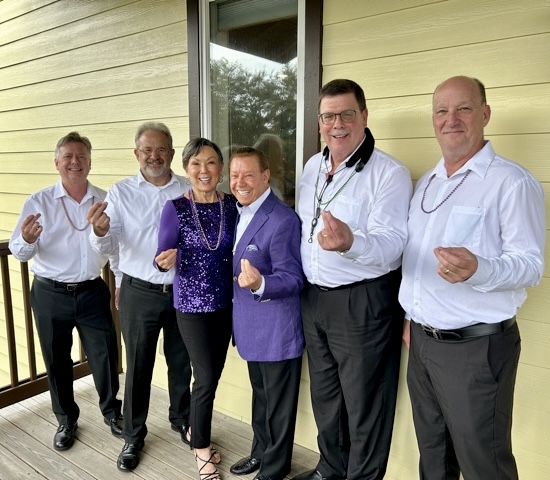 Six adults stand on a wooden porch, smiling and making small heart gestures with their fingers. Most wear white shirts; one woman and a man wear purple. They appear cheerful and festive.