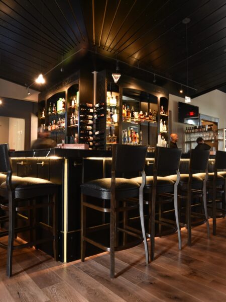 A modern bar with high black chairs, illuminated shelves stocked with liquor bottles, and warm lighting. The bar has a sleek black ceiling and wooden floor, creating an inviting atmosphere.
