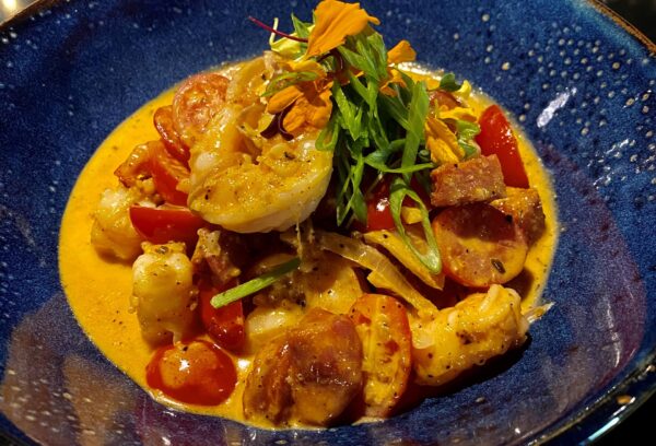 A blue bowl filled with creamy, orange-hued seafood stew, featuring shrimp, sausage, cherry tomatoes, herbs, and edible flowers, garnished with fresh greens.