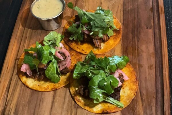 Three small tacos topped with greens, herbs, and pickled onions on a wooden board, served with a side of creamy sauce in a metal cup.