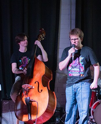 Two people perform on stage; one plays an upright bass and the other sings into a microphone. Both wear black t-shirts, and there is a drum set in the background.