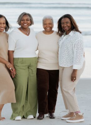 Four women stand side by side, smiling on a sandy beach with gentle ocean waves in the background. They are dressed in neutral tones and appear to be enjoying a relaxed, pleasant moment together.