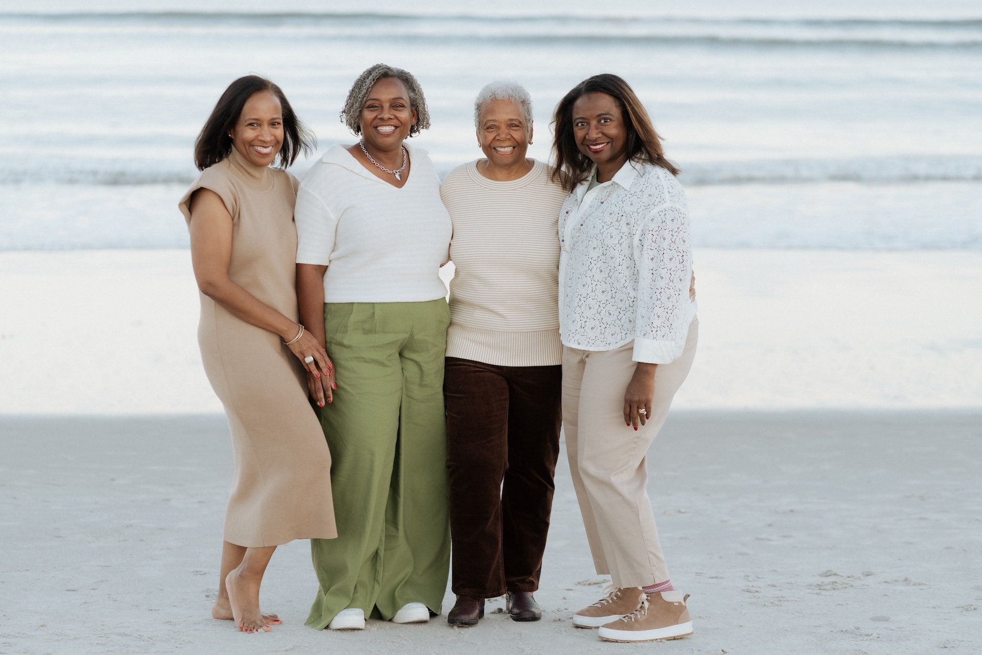 Four women stand side by side, smiling on a sandy beach with gentle ocean waves in the background. They are dressed in neutral tones and appear to be enjoying a relaxed, pleasant moment together.