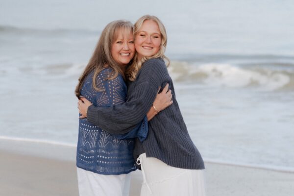 Two women smiling and hugging while standing on a sandy beach, with gentle ocean waves in the background. Both are wearing sweaters and light-colored skirts, enjoying a peaceful moment by the water.