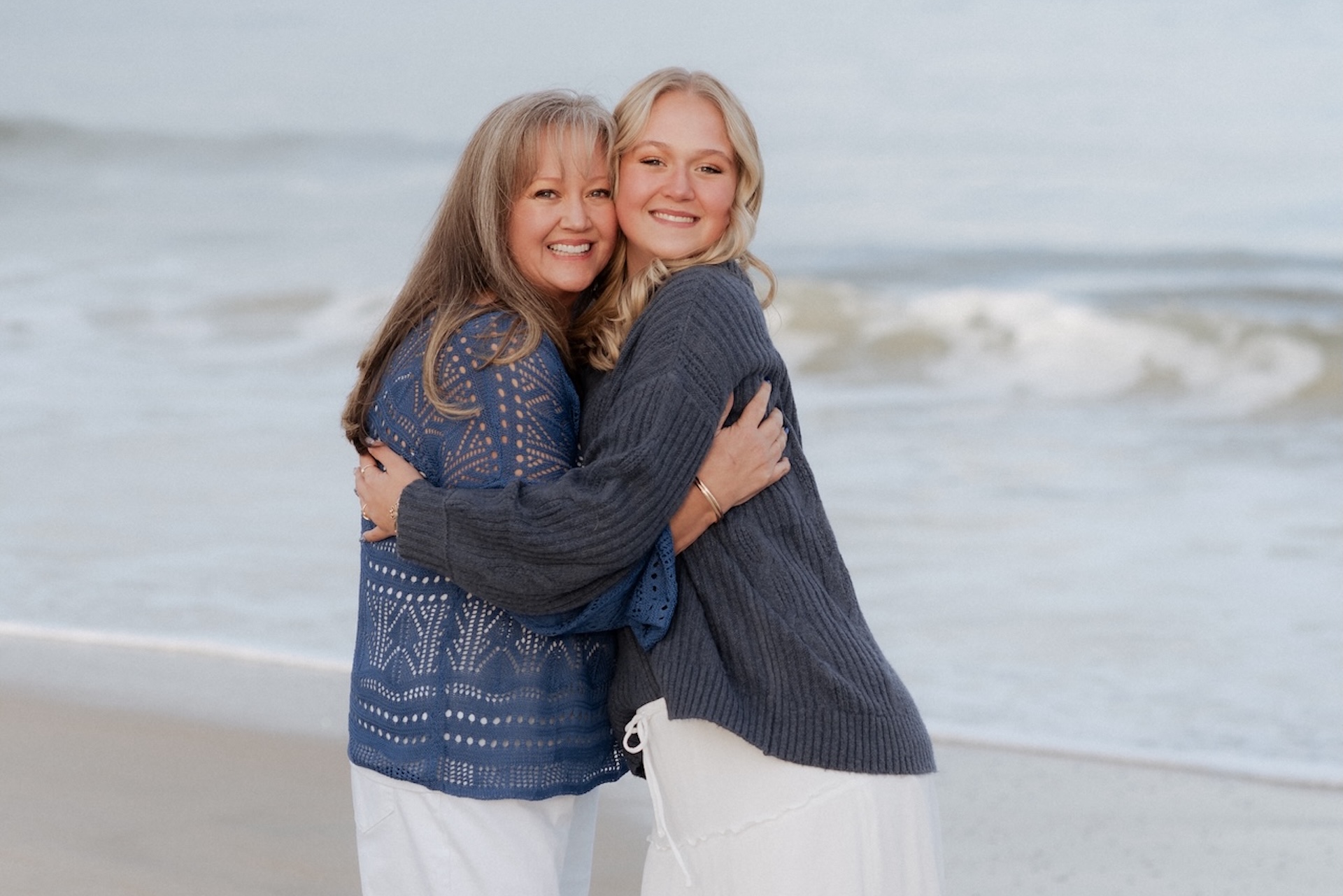 Two women smiling and hugging while standing on a sandy beach, with gentle ocean waves in the background. Both are wearing sweaters and light-colored skirts, enjoying a peaceful moment by the water.