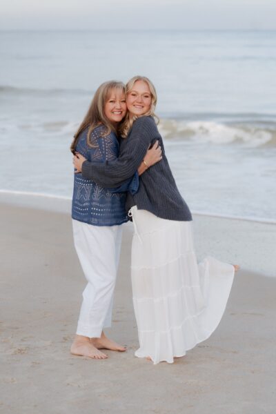 Two women stand barefoot on a sandy beach, smiling and hugging each other. Both wear sweaters and light-colored pants or skirts. Gentle ocean waves are visible in the background.