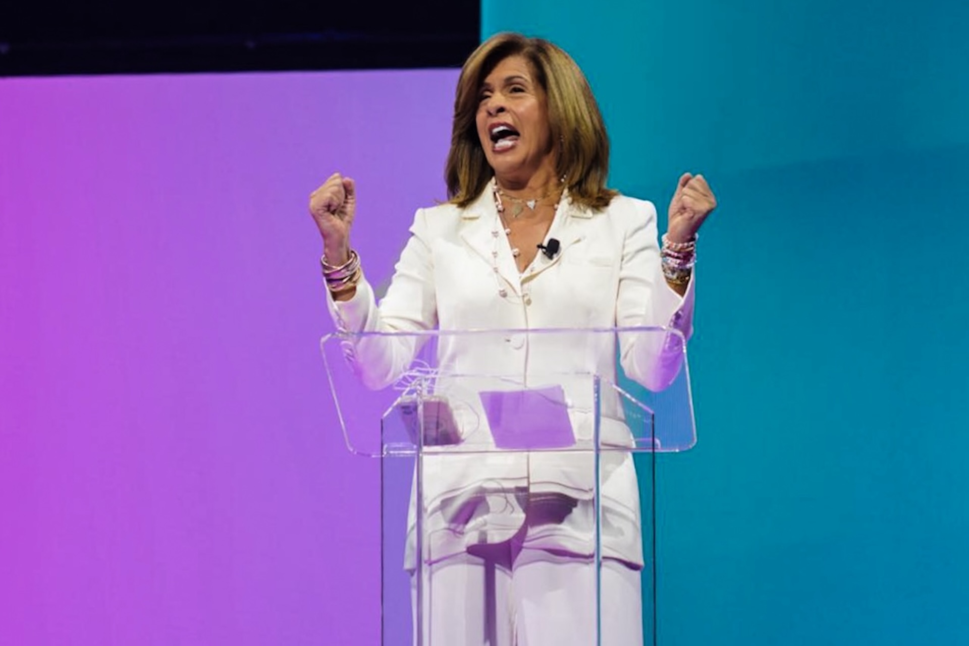 A woman in a white suit stands behind a clear podium, speaking passionately with her fists raised, against a purple and teal background.
