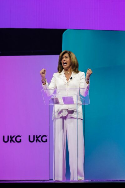 A woman in a white suit speaks energetically at a clear podium on a stage with purple and teal lighting and UKG logos displayed on the background wall.