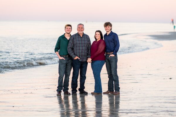 A family of four, two adults and two young men, stand together smiling on a sandy beach at sunset with gentle waves and a calm sea in the background.