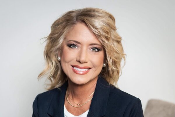 A woman with wavy blonde hair, wearing a dark blazer, white top, pearl earrings, and a gold necklace, smiles at the camera against a plain light background.