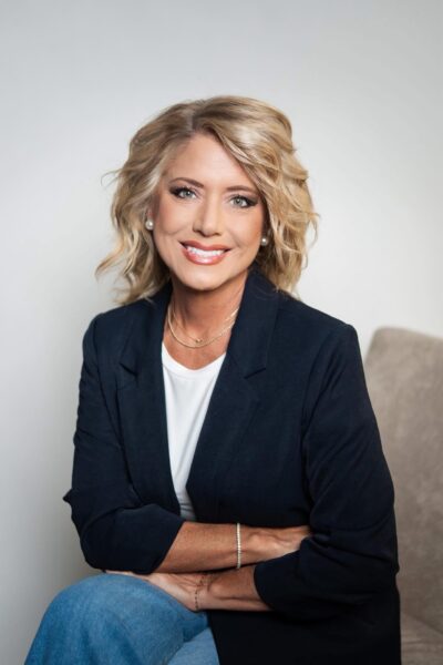 A woman with blonde, wavy hair sits smiling. She wears a navy blazer over a white top, blue jeans, and delicate jewelry. She poses with arms crossed, sitting on a beige chair against a light background.