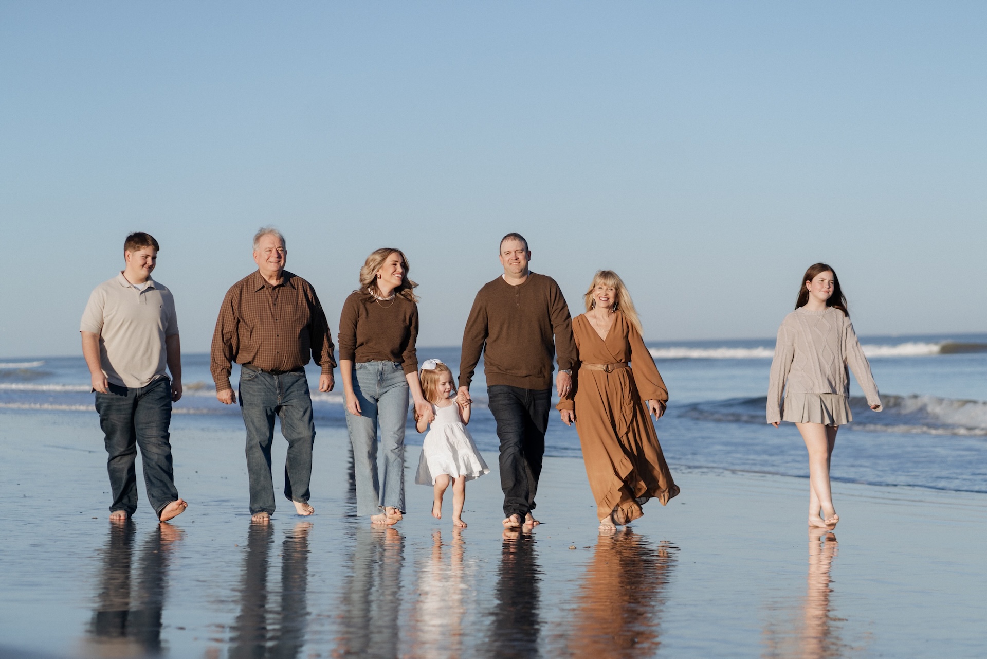 A group of seven people, including a child, walk barefoot along a beach shoreline on a sunny day, smiling and holding hands. The sky is clear and the ocean waves are gently rolling in.