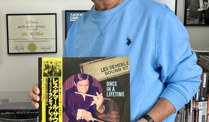 A smiling man in a blue sweatshirt stands in front of a wall with framed records and photos, holding a vinyl record titled Once in a Lifetime by Les Demerle Sound 67. Shelves of records line the background.