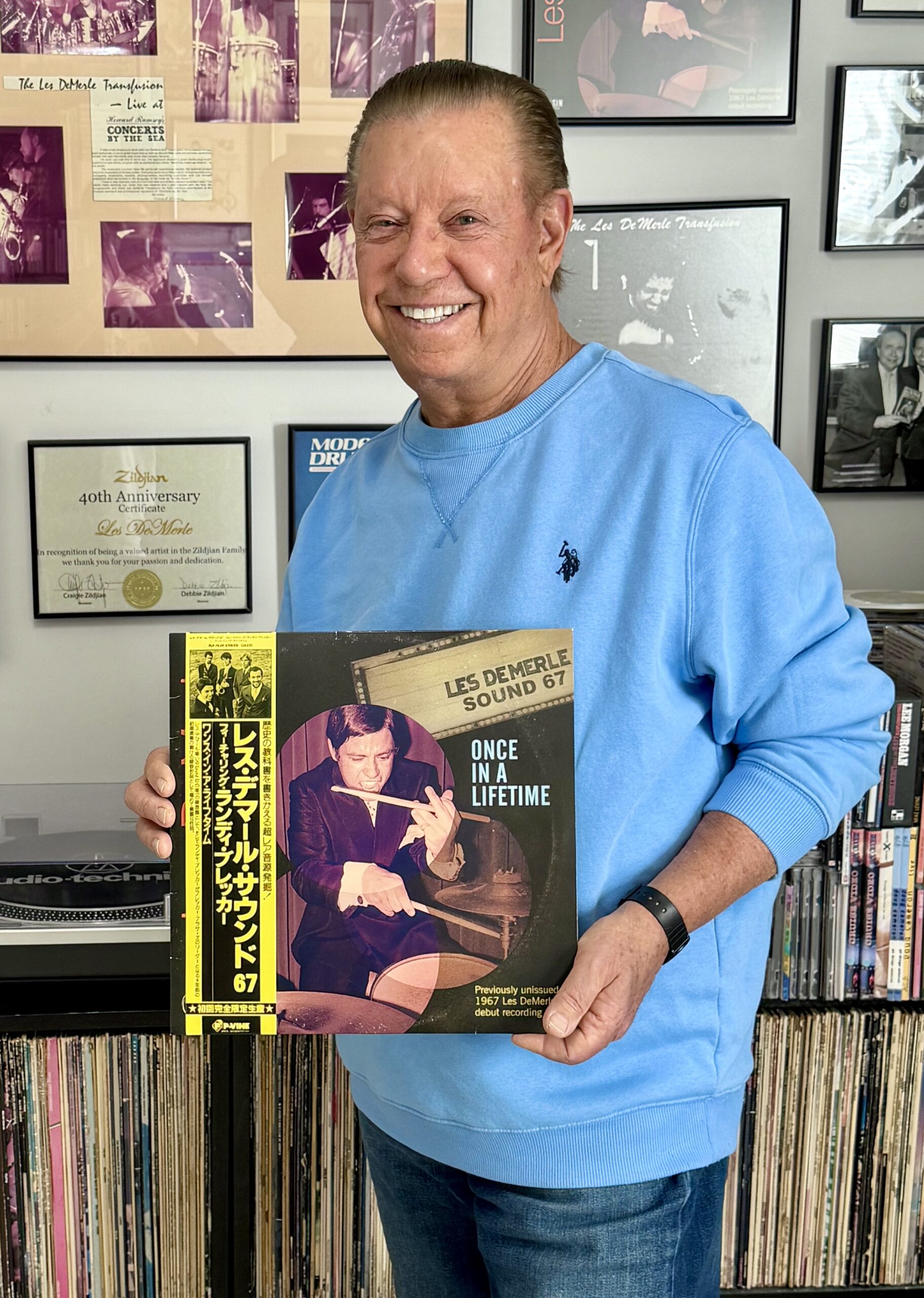 A smiling man in a blue sweatshirt stands in front of a wall with framed records and photos, holding a vinyl record titled Once in a Lifetime by Les Demerle Sound 67. Shelves of records line the background.