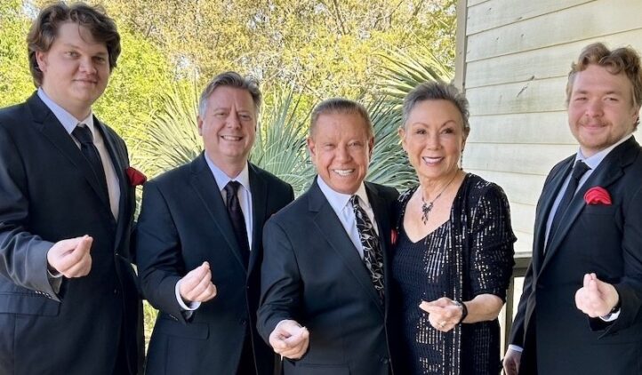 Five people dressed in formal attire stand outside, smiling and making small heart signs with their fingers. Trees and greenery are visible in the background next to a light-colored building.