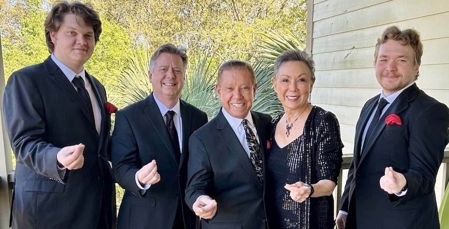 Five people dressed in formal attire stand outside, smiling and making small heart signs with their fingers. Trees and greenery are visible in the background next to a light-colored building.