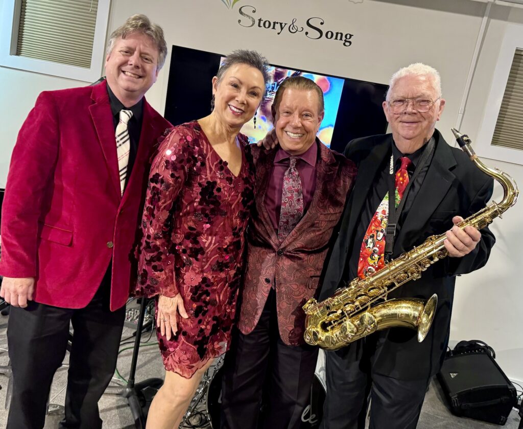 Four smiling adults, dressed in festive red and maroon attire, pose together indoors. One man holds a saxophone. A Story & Song sign and a TV screen are visible in the background.