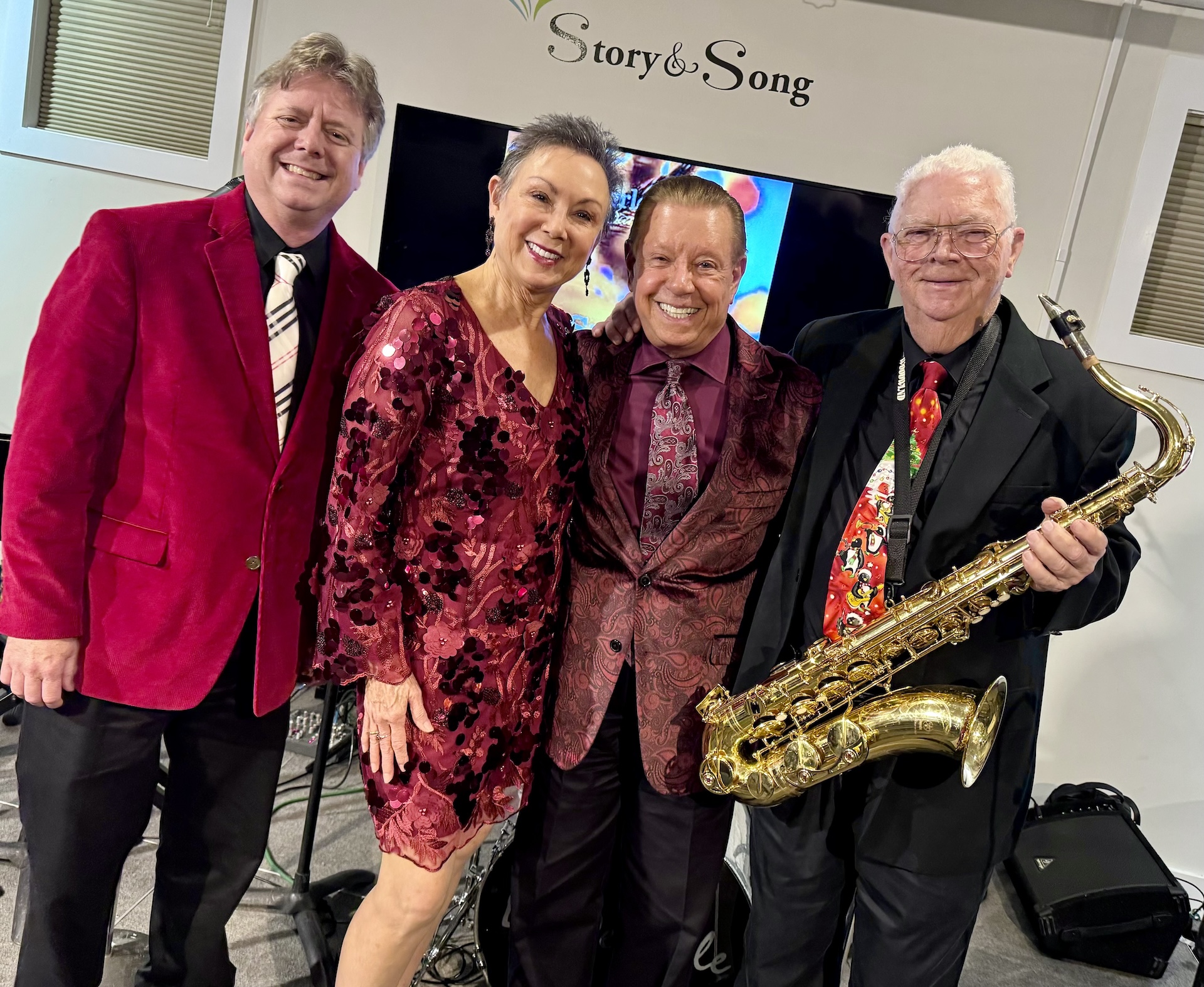 Four smiling adults, dressed in festive red and maroon attire, pose together indoors. One man holds a saxophone. A Story & Song sign and a TV screen are visible in the background.