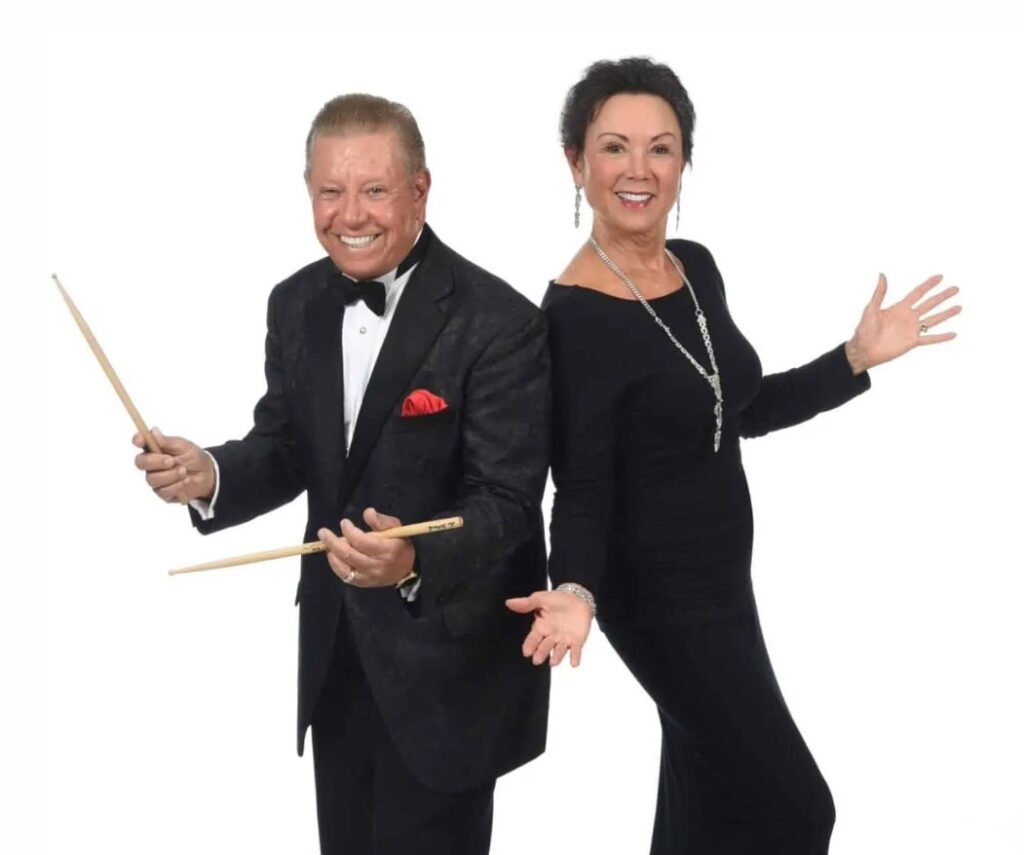 A smiling man in a tuxedo holds drumsticks next to a smiling woman in a long black dress, both posing enthusiastically against a plain white background.
