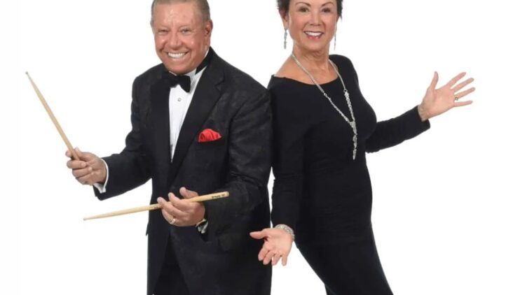 A smiling man in a tuxedo holds drumsticks next to a smiling woman in a long black dress, both posing enthusiastically against a plain white background.