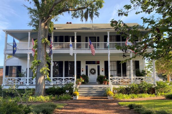 A two-story white house with black shutters, a wide front porch, and decorative columns. Multiple flags hang from the porch, and potted plants line the entrance. Trees and greenery surround the brick walkway to the door.