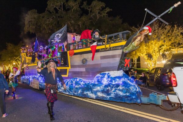 A festive pirate ship parade float decorated with lights and a mermaid figure moves down a street at night, with people dressed as pirates and illuminated trees in the background.