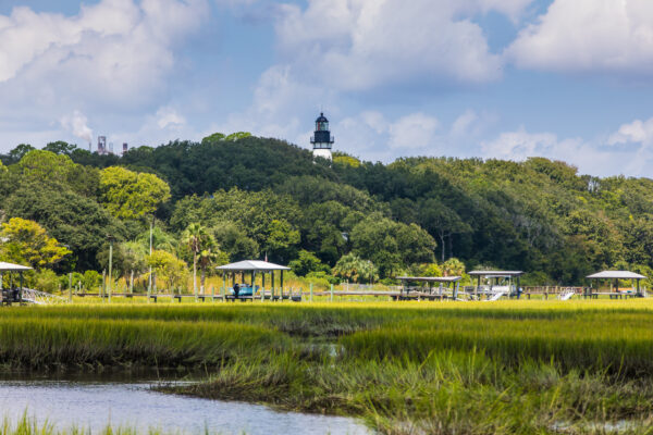 A lighthouse stands atop a tree-covered hill, with docks, shelters, and marsh grasses in the foreground under a partly cloudy sky.