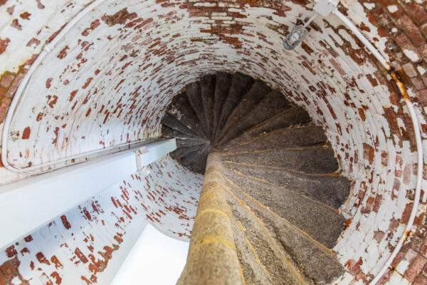 Aerial view looking down a narrow, spiral staircase made of worn stone steps encircled by a whitewashed, brick wall with patches of exposed red brick. A metal handrail follows the curve of the stairs.