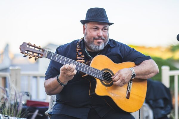 A man with a beard and hat sits outdoors playing an acoustic guitar, smiling and looking to the side. He wears a dark shirt and silver watch, with blurred background scenery.