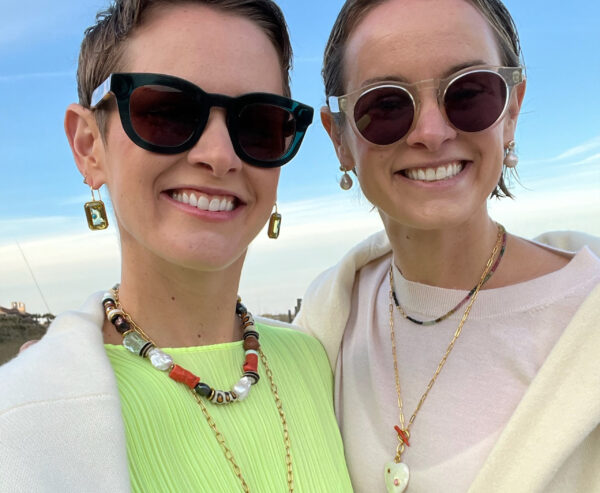 Two people are smiling outdoors, both wearing sunglasses, light-colored tops, gold jewelry, and earrings. They stand close together, with blue sky and blurred scenery in the background.