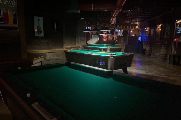 A dimly lit bar with two pool tables, one in the foreground and the other illuminated in the background. Barrels, wall art, and neon lights decorate the rustic interior.