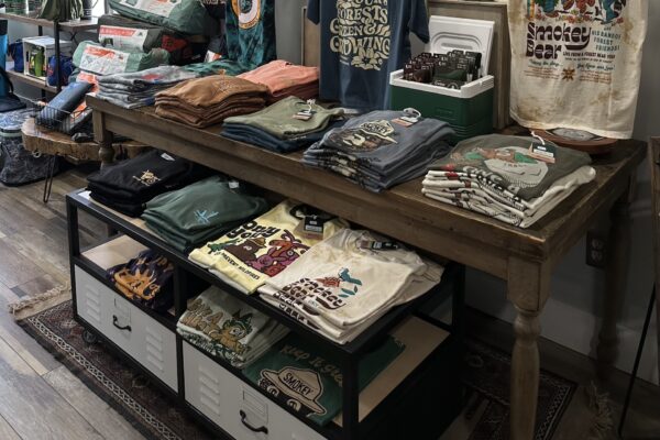 A display table in a store with neatly folded graphic t-shirts, some with nature and animal designs. More shirts are stored on the lower shelf and drawers underneath, all on a wooden floor with a patterned rug.