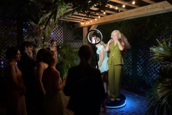 A woman in a green suit stands on a small platform under string lights, posing with another woman for a photo using a ring light, while a group of people in formal attire watch and smile at an outdoor nighttime event.