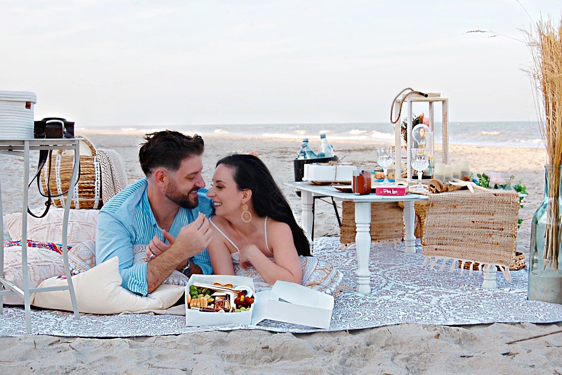 A couple lies on a blanket at the beach, smiling and holding hands, with a picnic setup including food, drinks, and decor on a low table beside them. The ocean and sandy shore are visible in the background.
